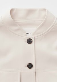 Crèmekleurig jack met een ronde kraag en twee donkere knopen aan de voorkant, met zichtbare stiknaden op zachte stof.