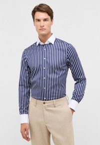 Navy gestreiftes Button-up-Hemd mit weißen Akzenten, langen Ärmeln und einem weißen Kragen; kombiniert mit beigen strukturierten Hosen.