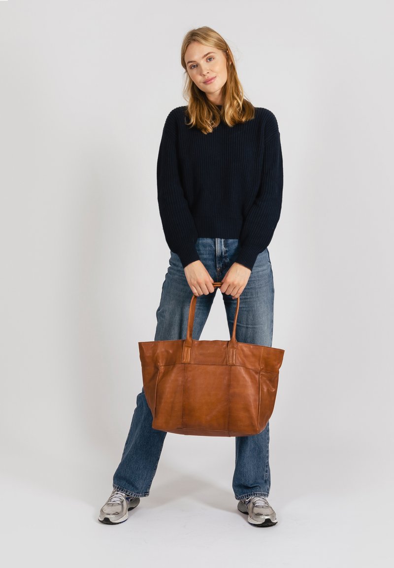 Brown leather tote bag with two handles, smooth texture, rectangular shape, and seams for structure. Worn by a person in casual attire.
