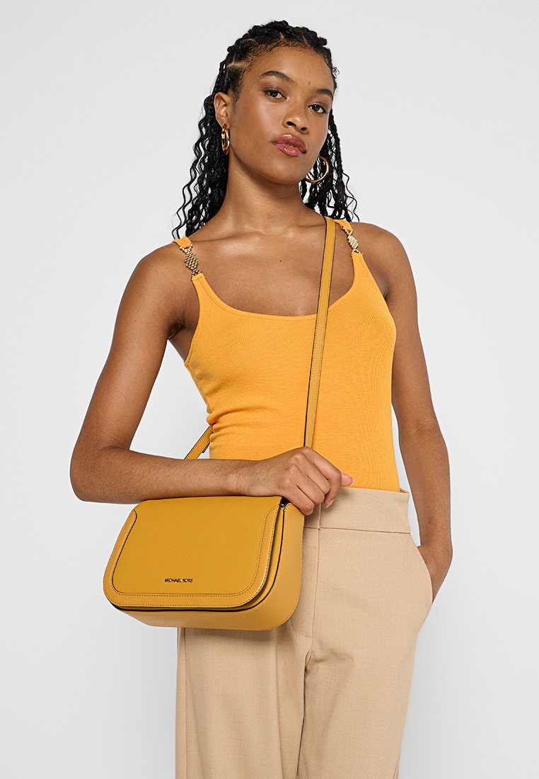 Frau mit geflochtenem Haar, die ein orangefarbenes Tanktop und beigefarbene Hosen trägt und eine senfgelbe Umhängetasche über der Schulter hat.