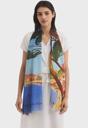 Kobieta w białej sukience, nosząca kolorowy szalik, na tle nadmorskiego krajobrazu z drzewami, wodą, budynkami i ludźmi na plaży.