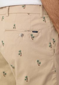 Tan cotton shorts with embroidered green palm trees, featuring a back pocket with a button closure and a branded label.