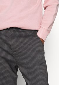 Person wearing dark gray pinstripe trousers with a hand in pocket and a light pink long-sleeve top.