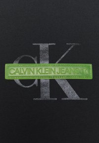 Czarna tkanina z dużym metalicznym srebrnym logo "K", z jasną zieloną prostokątną metką wytłoczoną napisem "CALVIN KLEIN JEANS".