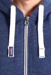 Sudadera con cremallera en azul marino con cuerda blanca. Cuenta con una cremallera plateada y una etiqueta de marca. Tejido suave y texturizado con un patrón jaspeado.