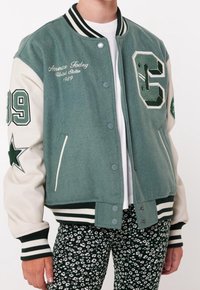 Green wool-blend varsity jacket with cream sleeves, ribbed cuffs, and hem. Features embroidered details, a star patch, and snap buttons.