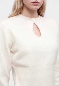 Jeune femme portant un pull crème avec un col en forme de trou de serrure fermé par un seul bouton, montrant une partie de son visage et de son buste.