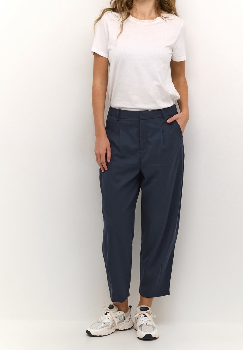 Femme portant un t-shirt blanc à manches courtes, un pantalon bleu marine taille haute et fuselé, et des baskets blanches, debout, une main dans la poche.