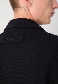 Chaqueta de HUGO en azul marino oscuro con textura acanalada, cuello con muescas y etiqueta de marca en la parte posterior. Cuenta con detalles de costura intrincados.