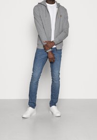 Grauer Zip-Hoodie mit Kordelzug, darunter ein weißes T-Shirt, blaue Jeans und weiße Sneakers. Hat ein kleines Logo auf der Brust.
