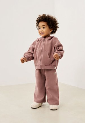 Kleinkind mit lockigem Haar trägt einen mauvefarbenen Kapuzenpullover mit Reißverschluss, passende Hose und weiße flauschige Hausschuhe, steht auf einem beigen Boden.