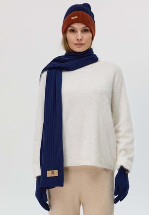 Marineblau gerippter Schal, orangefarbener gerippter Beanie, cremefarbenes, kuscheliges Sweatshirt, beigen Hosen und blauen Handschuhen mit einem Logotag. Weiche, strukturierte Materialien.