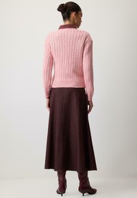 Žena s vlasmi v drdole, oblečená v ružovom svetríku s vrkočovým vzorom, dlhú tmavohnedú sukňu a bordové kozačky na opätku, stojaca pred jednoduchým pozadím.