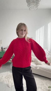 Donna che indossa un maglione rosso acceso a trecce e pantaloni neri, in piedi in una stanza luminosa con un divano bianco e un lampadario.