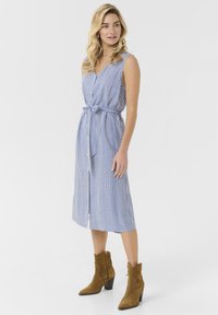 Vestido a rayas en azul y blanco, diseño sin mangas con escote en V y cintura ceñida con cinturón. Largo hasta medio pantorrilla, combinado con botas de tobillo de ante marrón.
