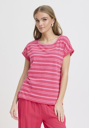 Jonge vrouw die een roze gestreept shirt met korte mouwen en effen roze broek draagt, staande tegen een effen witte achtergrond.