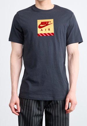 Granatowa bawełniana koszulka z złotym i czerwonym logo Nike z tekstem "AIR" w kwadracie, zestawiona z czarno-białymi spodniami w paski.