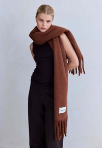 Bruine wollen sjaal met franjes, met een rechthoekig label met tekst. Het model draagt een zwarte tanktop en broek.