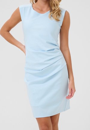 Femme portant une robe bleu clair sans manches, froncée, longueur genoux, posant avec une main sur la hanche devant un fond uni.