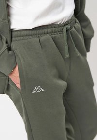 Persona che indossa pantaloni della tuta verde oliva con coulisse in vita e un logo con due figure sedute vicino alla tasca laterale, mano parzialmente inserita nella tasca.