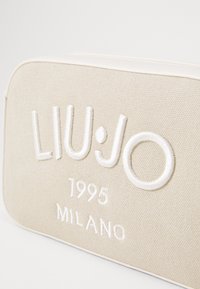 Borsa cosmetica in tela beige con ricamo bianco del testo "LIU·JO 1995 MILANO". Forma rettangolare con bordi lisci e una sottile texture.