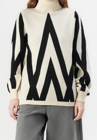 Pull col roulé crème avec des motifs zigzag noirs audacieux sur le devant et les manches, associé à un pantalon gris ample.