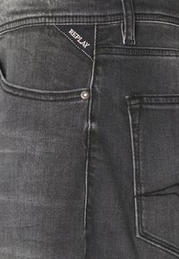 Jeans negre din denim cu un finisaj texturat, având un buzunar pe spate accentuat de o etichetă neagră cu logo-ul alb "REPLAY" și detalii cu rivete argintii.