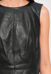 Femme portant un top en cuir noir sans manches avec des coutures visibles à l’avant et une texture lisse sur fond clair.
