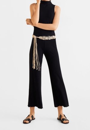 Czarny dzianinowy kombinezon z wysokim kołnierzem, szerokimi, skróconymi nogawkami, falistym dołem i misternym beżowym paskiem. Uzupełniony płaskimi czarnymi sandałami.