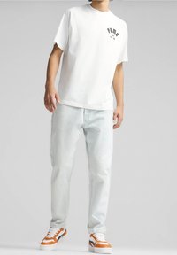 Camiseta blanca de algodón con el logo negro de Puma, jeans de mezclilla azul claro y zapatillas naranjas y blancas con acentos negros. Corte casual, mangas cortas.