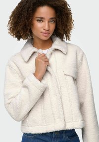 Giovane donna con capelli ricci che indossa una giacca di pile color crema e jeans blu, mentre tiene il colletto della giacca con una mano.