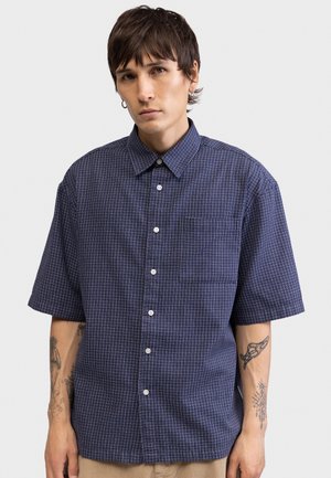 Fiatal személy rövid sötét hajjal, kék kockás rövid ujjú gombos ingben, mindkét alkarját látható tetoválások díszítik.