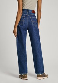 Jeans de denim azul de cintura alta e pernas largas, com uma textura leve, costuras contrastantes e dois bolsos traseiros. Acento de couro castanho na cintura.