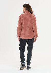 Fleece-pullovers i dammig ros med en texturerad vågig mönster; har en hög krage och långa ärmar, i kombination med svarta tapered byxor och vandringsskor.