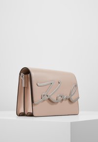 Beżowa prostokątna skórzana torba na ramię z srebrnym napisem "Karl" z przodu, stojąca na białej powierzchni na tle gładkiej ściany.