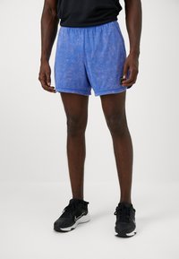 Blaue Sportshorts mit marmorierter Textur, ausgestattet mit einem elastischen Bund und einem kleinen Logo-Patch auf der linken Seite. Schwarze Sneakers werden getragen.