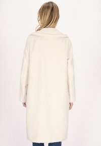 Cappotto lungo, bianco e fluffy con colletto a scialle e maniche lunghe, caratterizzato da una texture accogliente. Il cappotto arriva a metà polpaccio.