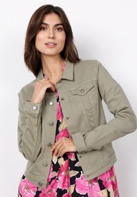 Olijfgroene denim jack met een knoopsluiting aan de voorkant, twee borstzakken en drukknoopafwerking bij de mouwen. Draag het over een bloemenjurk.