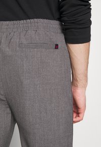 Grå byxor med elastisk midja, som har en enda bakficka med en liten röd och marinblå tag. Mjukt, strukturerat tyg med ett subtilt mönster.