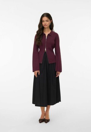 Cardigan bordeaux à fermeture éclair avec un col et des manches évasées, associé à une jupe noire longueur cheville et des chaussures plates marron.