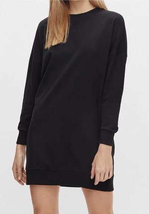 Kobieta ubrana w czarną, długorękawową, oversize'ową sukienkę-bluzę, stojąca na jednolitym jasnym tle, z rękami swobodnie opuszczonymi wzdłuż ciała.