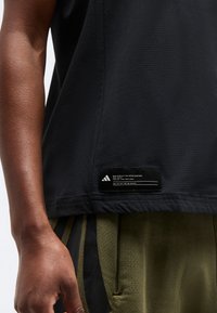 Κοντινή λήψη ενός ατόμου που φοράει μαύρο μπλουζάκι μπάσκετ Adidas με ετικέτα στο τελείωμα και χακί αθλητικές βερμούδες με μαύρες λωρίδες.