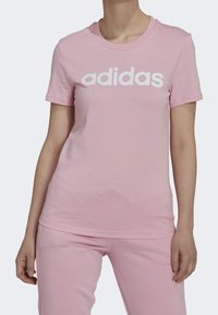 Růžové bavlněné tričko s krátkými rukávy, na přední straně s velkým bílým logem "adidas". Hladká textura, kulatý výstřih, volný střih.