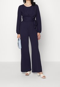 Marintröja med långa ärmar, invecklad design och knytning vid midjan. Modellen håller en ljusblå handväska och bär beige klacksandaler.