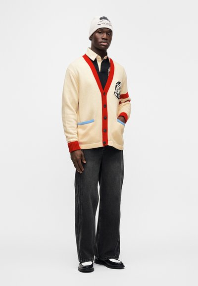 Κρεμ ζακέτα με κόκκινες λεπτομέρειες και μπλε περιμετρικά στο τσέπη, μαύρα κουμπιά και κεντημένο λογότυπο. Φορεμένη πάνω από μαύρο πουκάμισο και συνδυασμένη με γκρι τζιν.