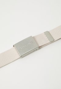 Lys beige vævet stofbælte med en sølvmetalspænde, der har mærkenavnet "JACK & JONES" præget. Enkel, rektangulær design.
