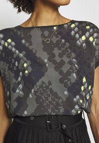 Blůza s krátkým rukávem z lehkého materiálu s tmavě šedým a žlutým vzorem. Kombinována s černou sukní s vysokým pasem a jednoduchým náhrdelníkem.