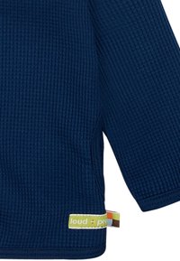 Maglietta a maniche lunghe blu scuro con una texture a coste e tasca laterale. Presenta un'etichetta verde e gialla "loud + proud" sull'orlo.