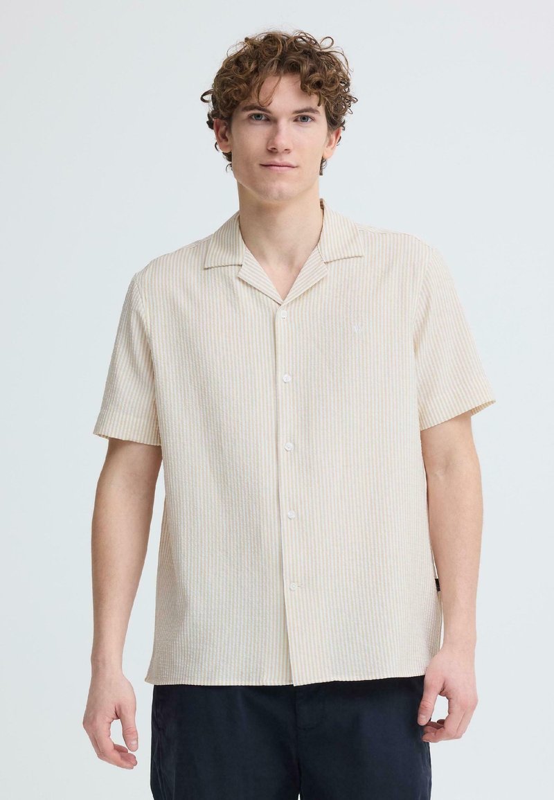 Jeune homme aux cheveux bouclés portant une chemise à manches courtes beige et blanche à rayures et un pantalon foncé, sur un fond clair uni.