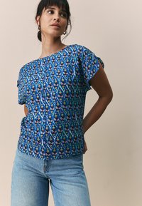 Haut bleu à motifs présentant des manches courtes, un col rond et un design floral en rouge, vert et jaune sur fond bleu.